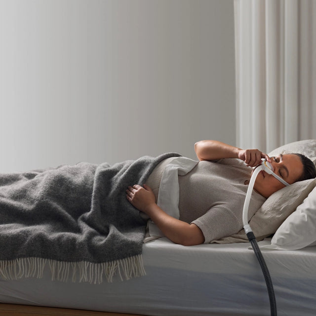 Woman lying in bed using a Fisher & Paykel Solo Nasal CPAP Mask with a gray blanket and white pillows.