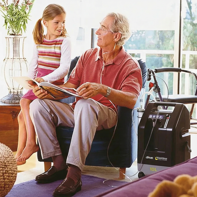 Man sitting in a chair with a young girl reading a book, with a DeVilbiss Compact 1025 10l/min Oxygen Concentrator on a table next to them.