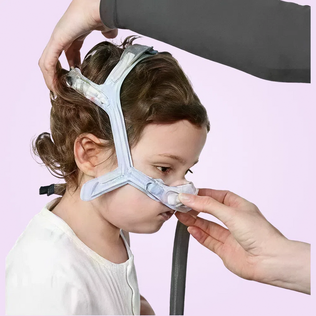 Person adjusting a ResMed Pixi Pediatric CPAP Mask on a child's face against a pink background