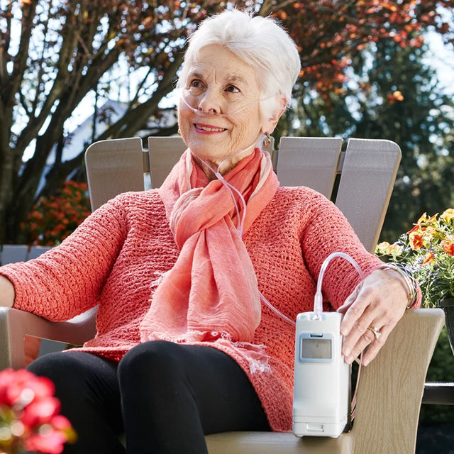 Senior woman sitting outdoors using Inogen One G4 System Portable Oxygen Concentrator, wearing a pink sweater and scarf.
