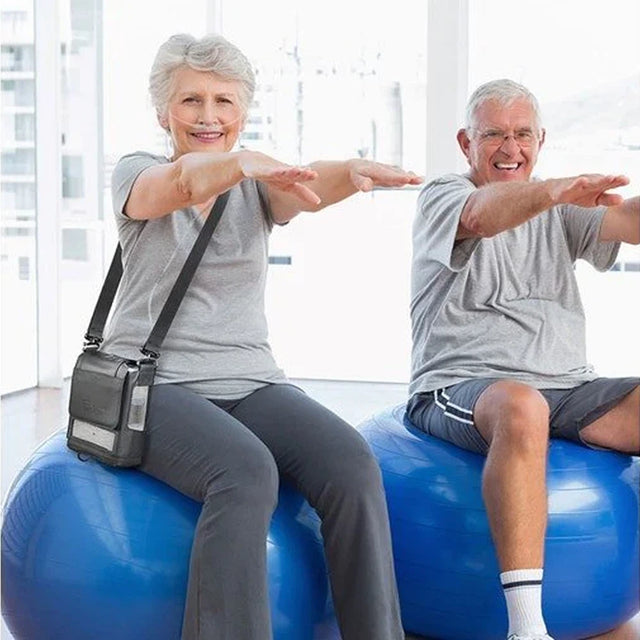 Two older adults exercising on blue fitness balls in a bright room while the woman is using Portable Oxygen Concentrator Inogen G5