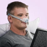 Man using a Fisher & Paykel Brevida Nasal Pillows CPAP Mask while reading a book with a pinkish-purple background