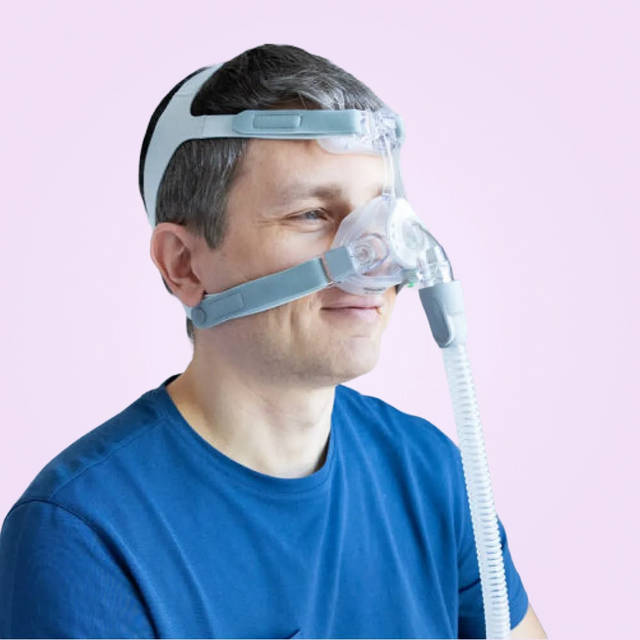 Man wearing a Löwenstein JOYCEone Nasal CPAP Mask with a clear hose on a pink background