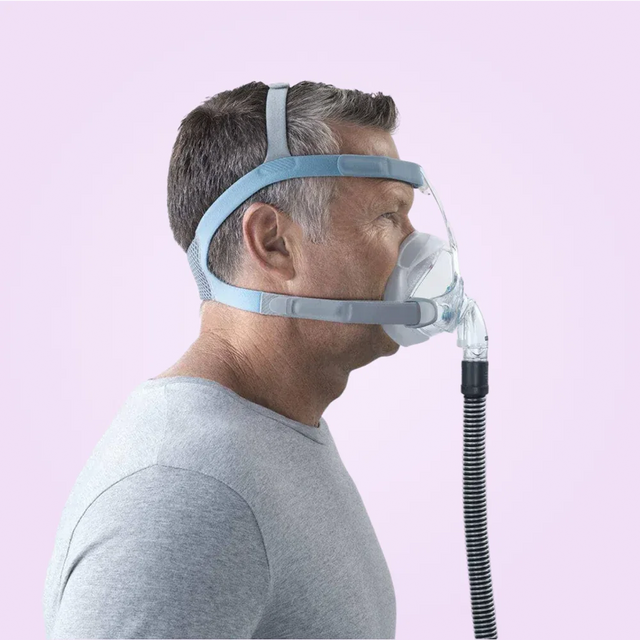 Person wearing a Fisher & Paykel Vitera Full Face CPAP Mask with a hose on a pink background
