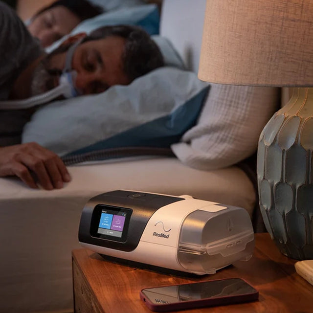ResMed AirSense 11 Autoset CPAP machine on a nightstand with a person using it in the background.