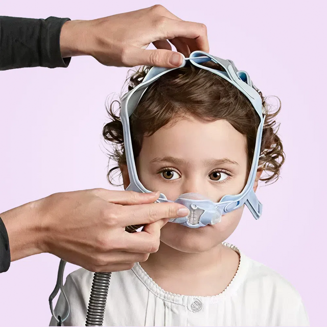 Child wearing a blue ResMed Pixi Pediatric CPAP Mask with a person adjusting it on a pink background