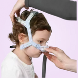 Person adjusting a ResMed Pixi Pediatric CPAP Mask on a child's face against a pink background