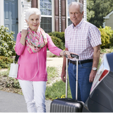 Senior couple with luggage standing outside a house, while the woman is using Inogen One G4 System Portable Oxygen Concentrator