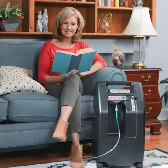 Woman reading a book on a couch next to a DeVilbiss 525 Oxygen Concentrator in a living room.