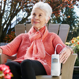Senior woman sitting outdoors using Inogen One G4 System Portable Oxygen Concentrator, wearing a pink sweater and scarf.