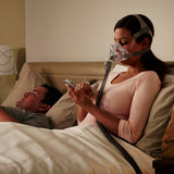 Woman using a smartphone with a man lying next to her in bed, while wearing ResMed AirFit™ F10 For Her Full Face Mask