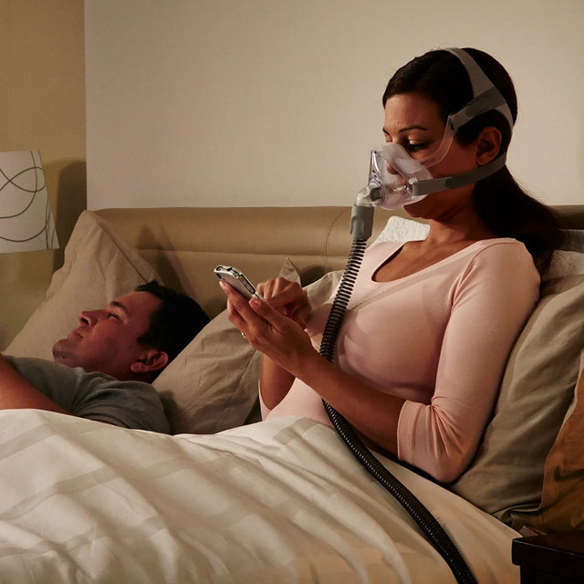 Woman using a smartphone with a man lying next to her in bed, while wearing ResMed AirFit™ F10 For Her Full Face Mask