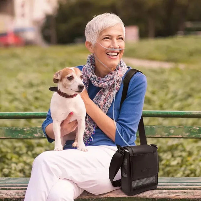 Woman sitting on a bench with a dog, smiling outdoors while using Portable Oxygen Concentrator Inogen G5